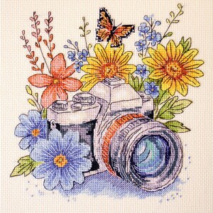 May include: Cross-stitch design of a camera surrounded by flowers and a butterfly. The flowers include yellow sunflowers, blue and orange blooms, and green leaves. The camera lens is detailed with shades of blue and grey.