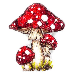 Op de afbeelding: Handgemaakte paddenstoelbroche met een grote rode kap met witte stippen, gemaakt van kralen. De steel is beige met bruin borduurwerk. Er zijn ook twee kleinere paddenstoelen, tegen een witte achtergrond.