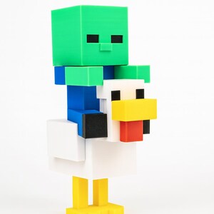 K&ouml;nnte beinhalten: Eine 3D-gedruckte Minecraft-Figur eines gr&uuml;nen Zombies, der auf einem wei&szlig;en Huhn reitet. Der Zombie hat schwarze Augen und sitzt auf einem Huhn mit gelbem Schnabel und F&uuml;&szlig;en. Die Figur besteht aus verschiedenfarbigen Bl&ouml;cken.