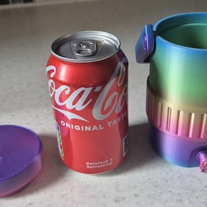 May include: A red Coca-Cola can next to a purple and blue ombre lid and a matching ombre drink holder. The drink holder has a tap and handles. The can reads "Coca-Cola Original Taste Delicious & Refreshing."