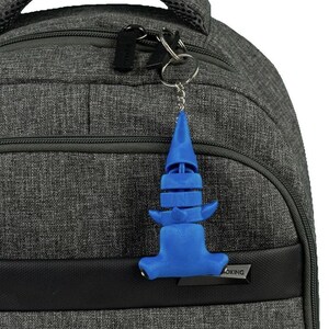 Könnte beinhalten: Ein blauer Plastik-Haifisch-Schlüsselanhänger, der an einem silbernen Schlüsselring hängt, der an einem grauen Rucksack befestigt ist.