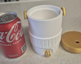 Könnte beinhalten: Ein weißer Getränkekühler mit goldenen Akzenten, einem passenden Deckel und einem Zapfhahn. Neben dem Kühler steht eine rote Coca-Cola-Dose. Auf der Dose stehen die Worte "ORIGINAL TASTE" und "Delicious & Refreshing".