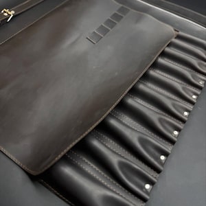 Può includere: Un astuccio portautensili in pelle marrone scuro con una grande patta, una serie di tasche verticali cucite per singoli oggetti e una chiusura con fibbia metallica. L'organizer include una sezione superiore con quattro piccole fessure.