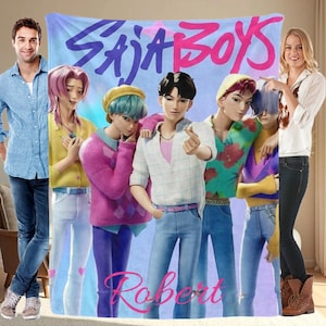 May include: A large, rectangular blanket with a group of stylized figures. The blanket has the words "SAJA BOYS" in a vibrant, multi-coloured font across the top, with the name "Robert" in a cursive font at the bottom. The background is a gradient of blue and purple.
