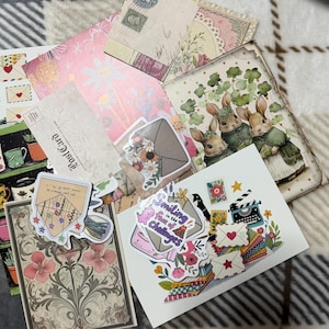 May include: A collection of decorative paper ephemera, including postcards, stickers, and cards. The items feature floral, bunny, and envelope designs. Colors include pink, green, and beige. Some items have text, including "Postcard" and "Smiling in the face of Challenges."