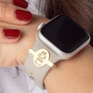 May include: A gold-toned watch band accessory with a paw print design and the word "Oscar" engraved on it. The accessory is attached to a gray Apple Watch.