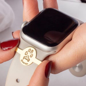 May include: A beige Apple Watch band with a gold paw print charm and the name "Oscar" engraved on it.