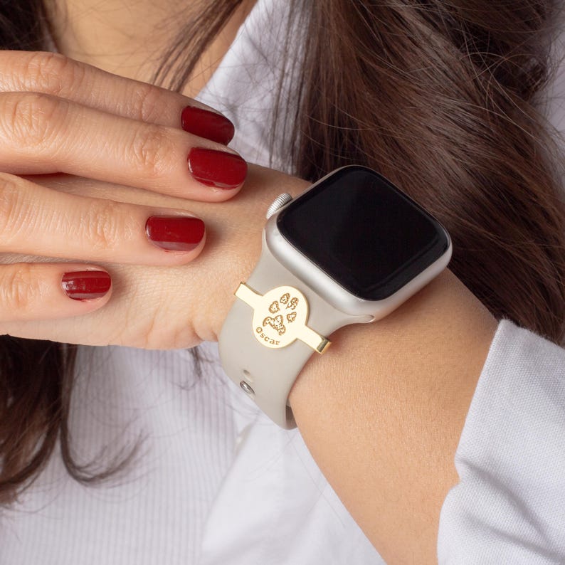 May include: A gold-toned watch band accessory with a paw print design and the word "Oscar" engraved on it. The accessory is attached to a gray Apple Watch.