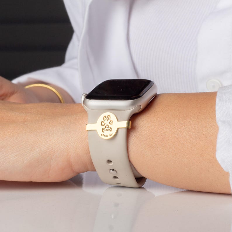 May include: A gold-colored watch band charm with a paw print and the name "Oscar" engraved on it. The charm is attached to a gray Apple Watch band.