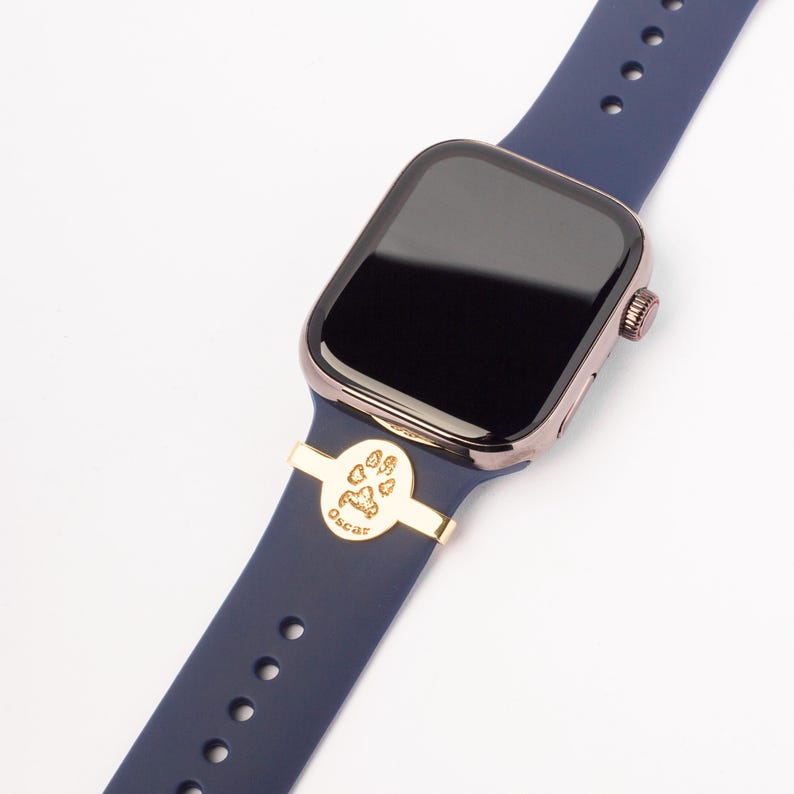 May include: A navy blue Apple Watch band with a gold-colored paw print charm that says "Oscar" on it.