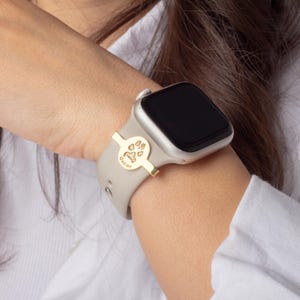 May include: A gold-colored watch band charm with a paw print and the name "Oscar" engraved on it. The charm is attached to a light gray Apple Watch band.