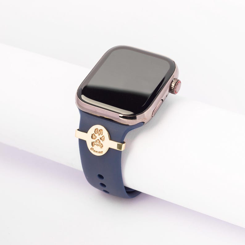 May include: A navy blue Apple Watch band with a gold paw print charm and the name "Oscar" engraved on it.