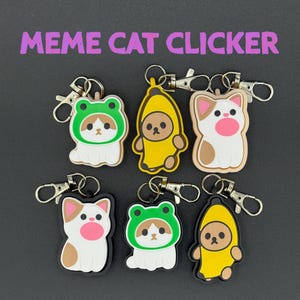 Op de afbeelding: Zes katvormige sleutelhangers met karabijnsluitingen. De sleutelhangers hebben kattenontwerpen verkleed als een kikker, een banaan en een kat. De tekst "Meme Cat Clicker" wordt in het paars weergegeven.