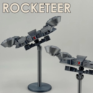 Może przedstawiać: Dwa szare, futurystyczne modele statku kosmicznego, każdy zamontowany na szarym stojaku. Modele mają złożony, geometryczny kształt z czerwonymi akcentami. Nad modelami wydrukowano słowo "ROCKETEER".