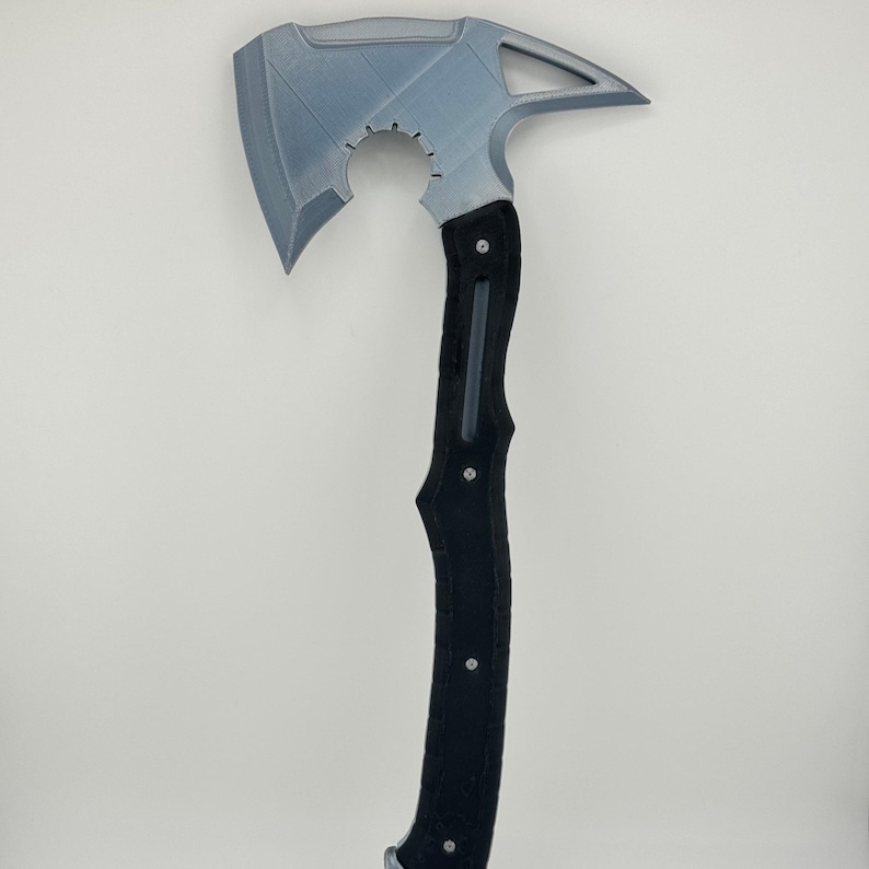 May include: A gray and black axe with a unique design. The axe head is gray with a curved blade and a pointed end. The handle is black with a textured grip and silver rivets. The axe has a hole near the top of the head.