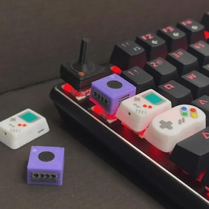May include: A mechanical keyboard with custom keycaps in the shape of retro video game consoles and controllers. Keycaps include a purple GameCube, a white Game Boy, and a joystick. The keyboard has red LED backlighting.