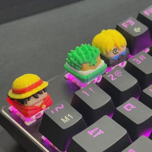 以下が含まれることがあります： アニメのキャラクターデザインが施されたカスタムキーキャップを備えたメカニカルキーボードのクローズアップ。キーキャップはカラフルで詳細で、赤、黄、緑のキャラクターがいます。キーボードは、紫色のバックライトを備えた黒いキーを備えています。