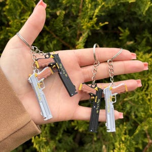 May include: Keychains featuring miniature gun replicas in silver and black, with gold and brown accents. Each keychain has a ring and chain for attachment. The text "Easy Say Gang" is visible on the black gun replicas.