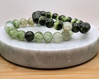 May include: Two beaded bracelets with green and white stones. The bracelets are made of round, polished beads in varying shades of green and white. The bracelets are displayed on a white, circular platform, set against a light background.