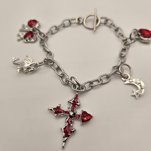 May include: Silver-toned charm bracelet featuring a cross with red enamel and a heart-shaped gem. Additional charms include a dragon, crescent moon, stars, and red heart-shaped gems. The bracelet has a toggle clasp.