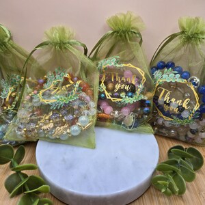 May include: A collection of beaded bracelets presented in small green mesh bags tied with green ribbons. Some bags feature a gold-framed "Thank you" tag. The bracelets display a variety of colours and bead sizes. The bags are arranged on a white marble stand.