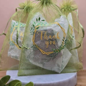 Puede incluir: Varias bolsas pequeñas de organza verde con cordones, cada una con un regalo. Las bolsas están decoradas con un diseño geométrico dorado y las palabras "Thank you" en letras doradas, rodeadas de hojas verdes.