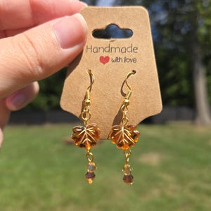May include: A pair of handmade gold-colored earrings with leaf-shaped charms and dangling beads. The earrings are displayed on a brown tag that reads "Handmade with love."