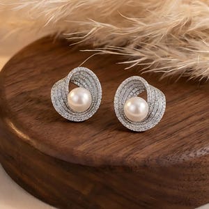 Peut inclure: Une paire de boucles d'oreilles à clous avec une perle centrale entourée d'un motif argenté tourbillonnant. Les boucles d'oreilles sont posées sur une surface en bois foncé, avec des plantes plumeuses séchées en arrière-plan.