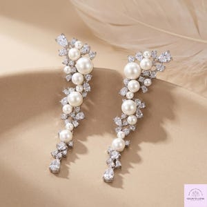 Può includere: Un paio di orecchini color argento. Ogni orecchino presenta un design a cascata di grandi perle bianche rotonde e cristalli trasparenti scintillanti. Gli orecchini hanno una forma a goccia, con un cristallo a forma di pera in basso.