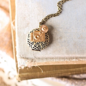 May include: A gold-toned camera locket necklace with a filigree design. The locket is open and the camera charm is visible. The necklace is on a brown book.