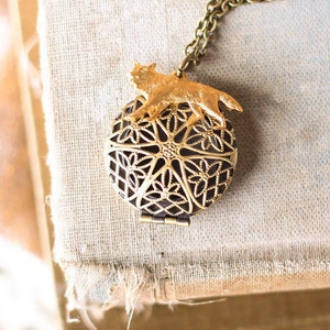 May include: A gold locket necklace with a detailed filigree design and a small gold animal figure on top. The locket is open and the inside is black.