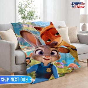 May include: A plush blanket featuring Judy Hopps and Nick Wilde from Zootopia. Judy, a gray rabbit, is giving a peace sign, while Nick, an orange fox, is smiling. The blanket is draped over a gray sofa. The text "SHIP US Now" is in the upper right corner.