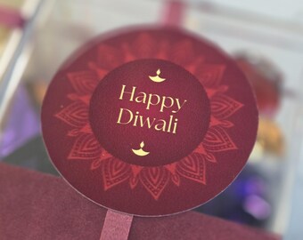 May include: A circular maroon sticker with a gold mandala pattern and the text Happy Diwali in gold lettering, featuring two small gold diya lamp icons above and below the text, attached to a clear box with a matching maroon ribbon.