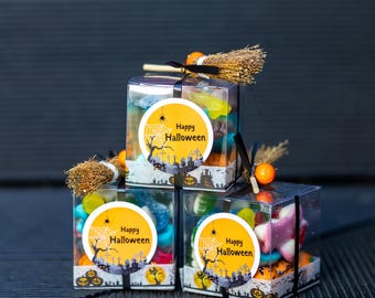 May include: Three clear square plastic boxes filled with assorted colorful candies. Each box features a round yellow sticker with a spiderweb, a spider, and the text Happy Halloween. A miniature straw broom is attached to the top of each box.