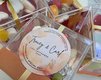 May include: Clear square plastic favor boxes filled with assorted colorful gummy candies and white heart-shaped marshmallows. Each box is wrapped with a gold ribbon and topped with a small fabric flower. A round sticker reads: Tracey & Carl, 11th September 2025, Thank you for joining us in sharing our special day.