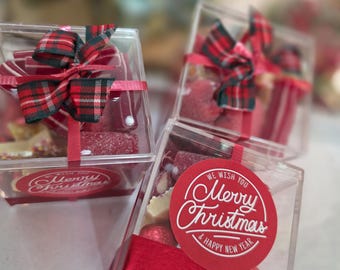 May include: Clear square boxes filled with assorted candies, tied with red ribbons and plaid bows. Each box has a red circular sticker with the text "Merry Christmas & Happy New Year". The boxes are arranged on a surface.
