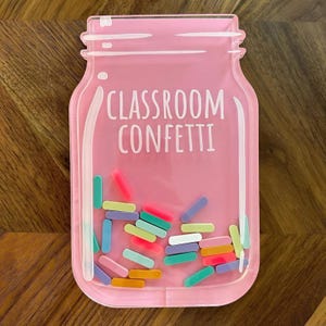 Könnte beinhalten: Ein rosa glasförmiger Behälter mit den weißen Worten "CLASSROOM CONFETTI". Der Behälter ist mit bunten, zylindrischen Konfetti-Stücken in verschiedenen Farben gefüllt, darunter Rosa, Blau, Grün und Gelb. Das Glas steht auf einer Holzoberfläche.