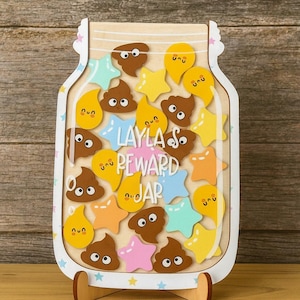 Peut inclure: Un pot de récompenses en bois en forme de bocal, rempli de jetons colorés en forme d'étoiles, de gouttes et de caca. Le pot est personnalisé avec le nom "Layla's" et les mots "Reward Jar". Le pot repose sur un support en bois.
