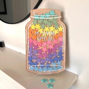以下が含まれることがあります： カラフルな星の切り抜きで満たされた瓶型の木製サイン。サインには白い文字で「MS CUMMINS SUPERSTARS」と書かれています。いくつかの緩い星が瓶の下に散らばっています。瓶は白い表面に表示されています。