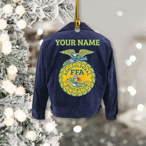 Op de afbeelding: Marineblau jasvormig ornament met de tekst "YOUR NAME" boven een geel en blauw FFA-embleem met een adelaar, uil en de woorden "Agricultural Education". De jas heeft een gouden ophanglus.