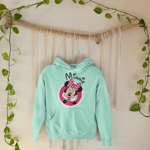May include: A light blue hoodie featuring a Minnie Mouse graphic. The graphic includes Minnie's face, a pink bow, and the name "Minnie" in a script font. The hoodie has a front pocket and a hood.
