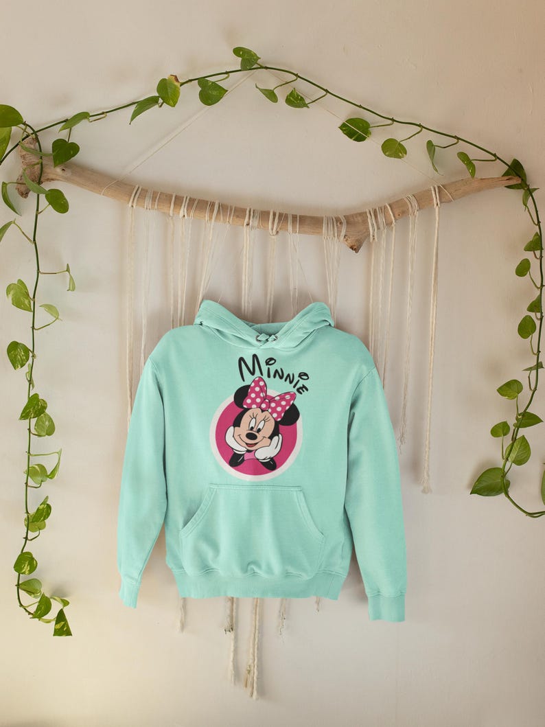 May include: A light blue hoodie featuring a Minnie Mouse graphic. The graphic includes Minnie's face, a pink bow, and the name "Minnie" in a script font. The hoodie has a front pocket and a hood.