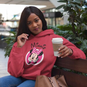 May include: A red Minnie Mouse hoodie with a white and pink graphic. The hoodie features a drawstring hood and long sleeves. The person in the image is holding a white coffee cup with a brown sleeve.
