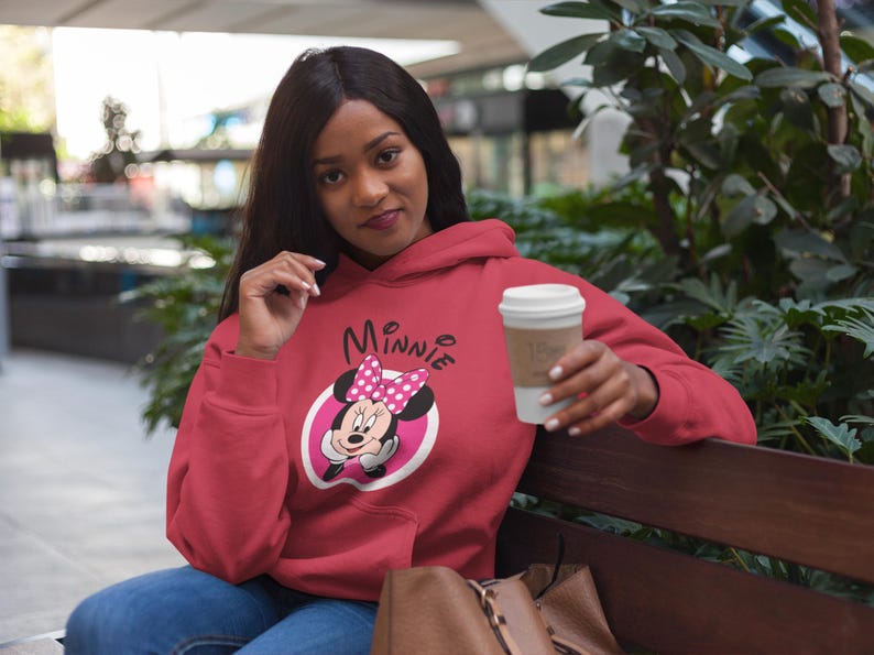 May include: A red Minnie Mouse hoodie with a white and pink graphic. The hoodie features a drawstring hood and long sleeves. The person in the image is holding a white coffee cup with a brown sleeve.