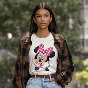 May include: White t-shirt featuring a Minnie Mouse graphic with a pink polka dot bow. The shirt is worn with a brown plaid shirt, blue jeans, and a brown belt. The background is a blurred outdoor scene.