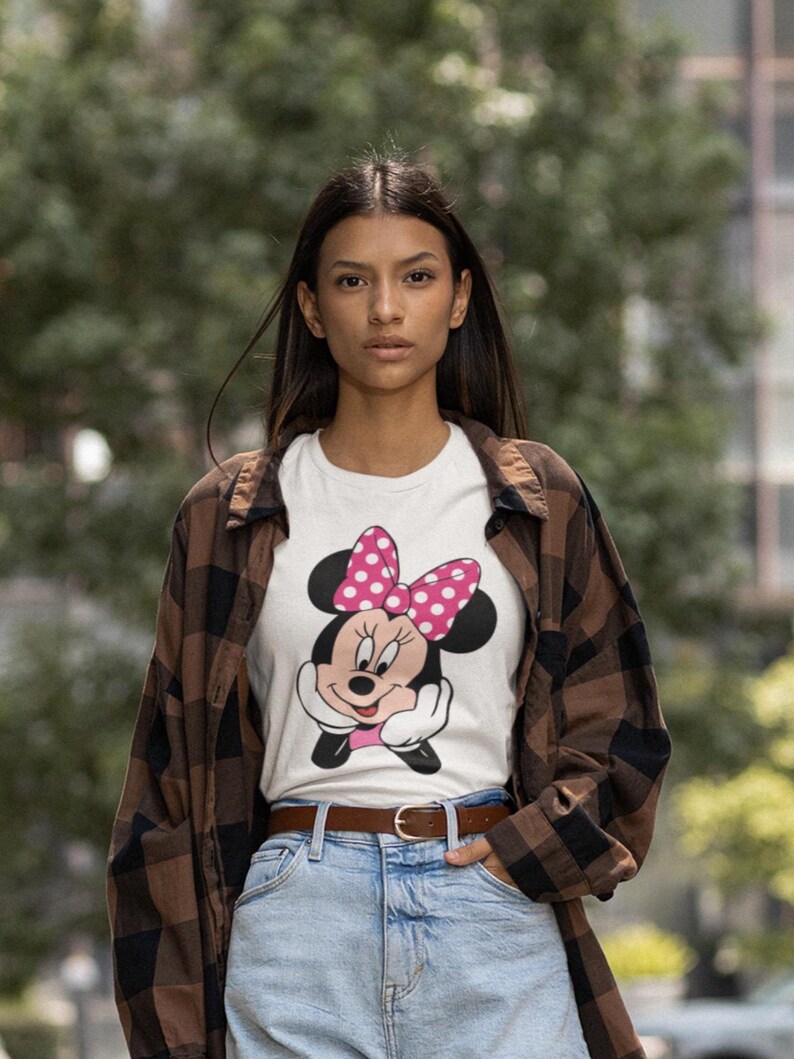 May include: White t-shirt featuring a Minnie Mouse graphic with a pink polka dot bow. The shirt is worn with a brown plaid shirt, blue jeans, and a brown belt. The background is a blurred outdoor scene.