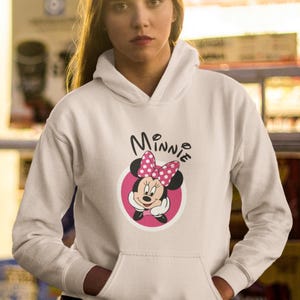 May include: White hooded sweatshirt featuring a Minnie Mouse graphic. The graphic includes Minnie's face, a pink bow with white polka dots, and the name "Minnie" in black script. The sweatshirt has a front pocket.