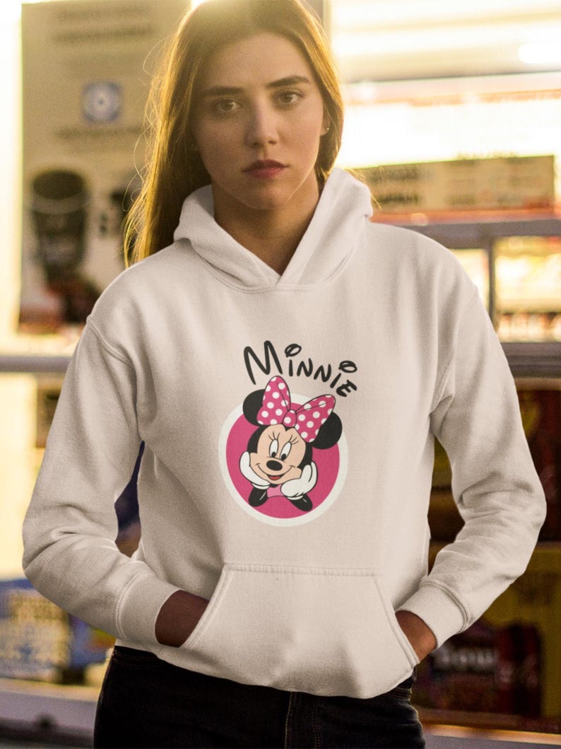 May include: White hooded sweatshirt featuring a Minnie Mouse graphic. The graphic includes Minnie's face, a pink bow with white polka dots, and the name "Minnie" in black script. The sweatshirt has a front pocket.