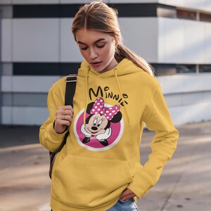 May include: Mustard yellow hoodie featuring a Minnie Mouse graphic. The graphic includes Minnie's face, a pink bow with white polka dots, and the word "Minnie" in black script. The hoodie has a kangaroo pocket and a drawstring hood.