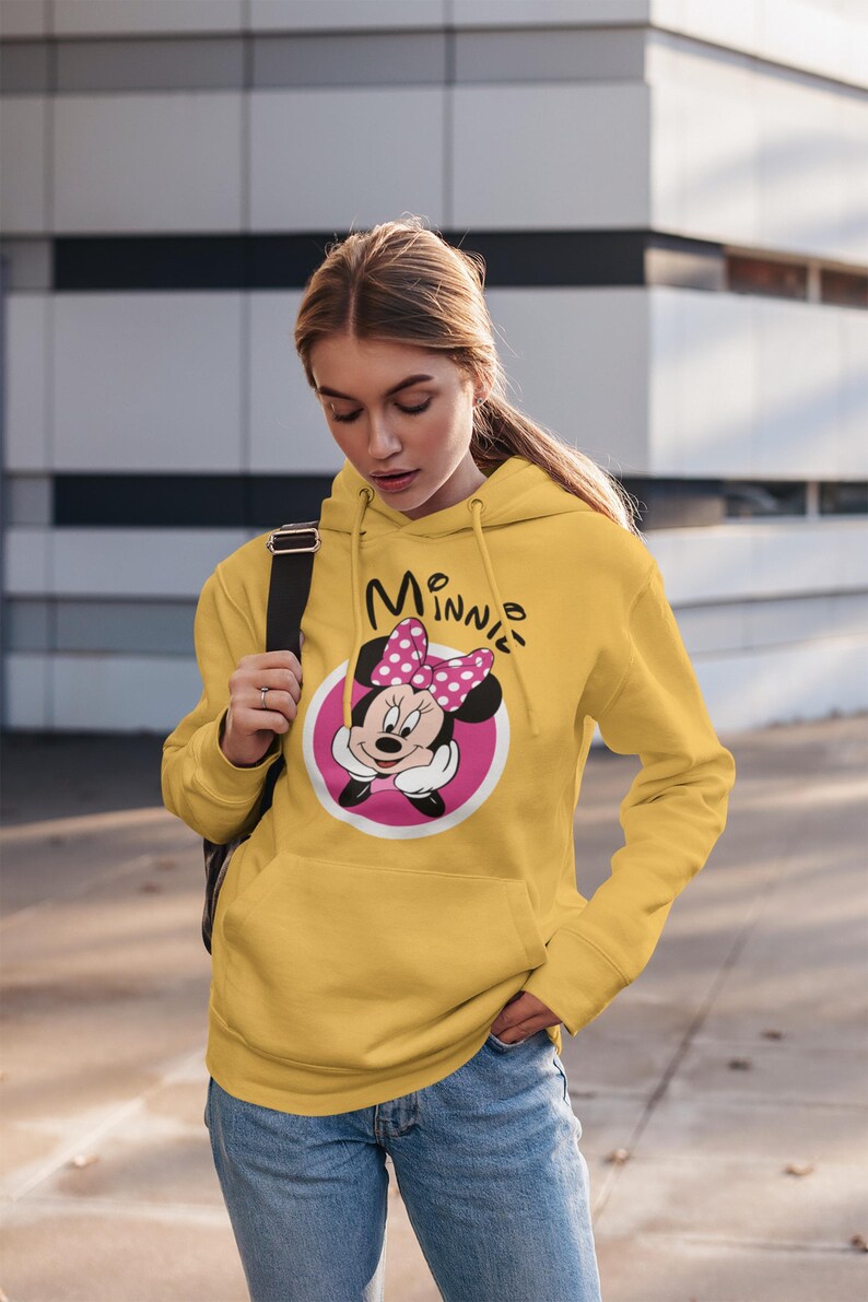 May include: Mustard yellow hoodie featuring a Minnie Mouse graphic. The graphic includes Minnie's face, a pink bow with white polka dots, and the word "Minnie" in black script. The hoodie has a kangaroo pocket and a drawstring hood.